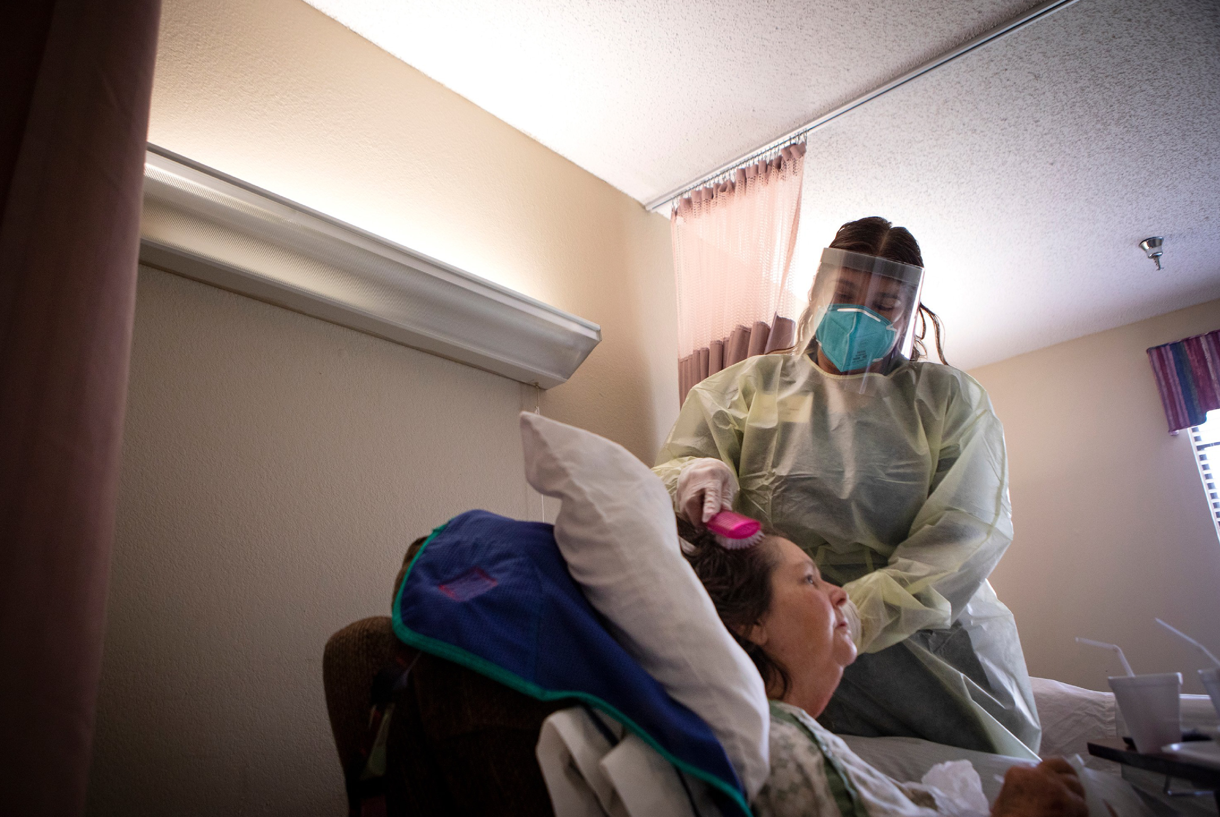 Photo of a nurse with mask and face shield combing a woman’s hair in a bed.